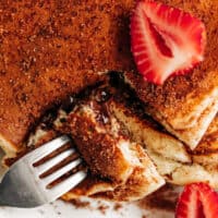 top view of a pancake stack, decorated in cinnamon sugar and fresh strawberries slice, fork opening a cut edge of stack to see the runny chocolate centre.