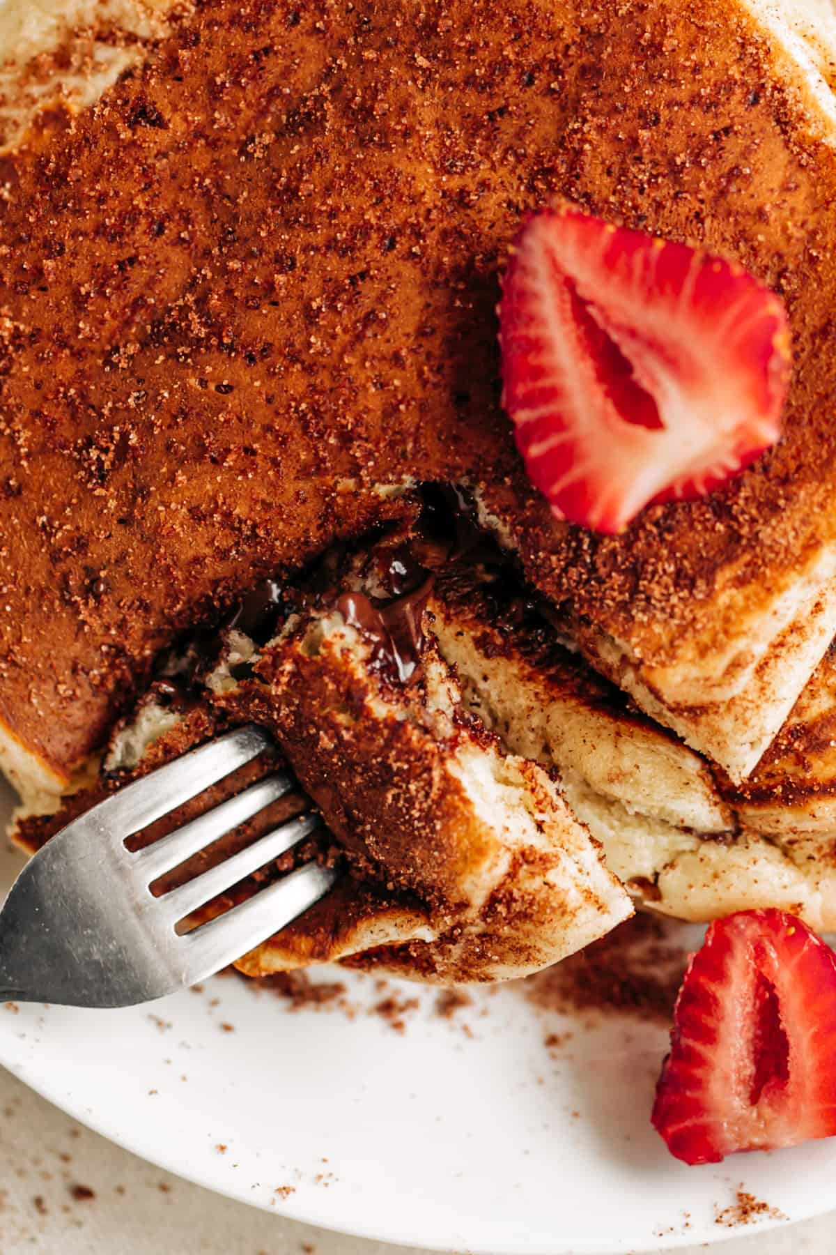 top view of a pancake stack, decorated in cinnamon sugar and fresh strawberries slice, fork opening a cut edge of stack to see the runny chocolate centre.