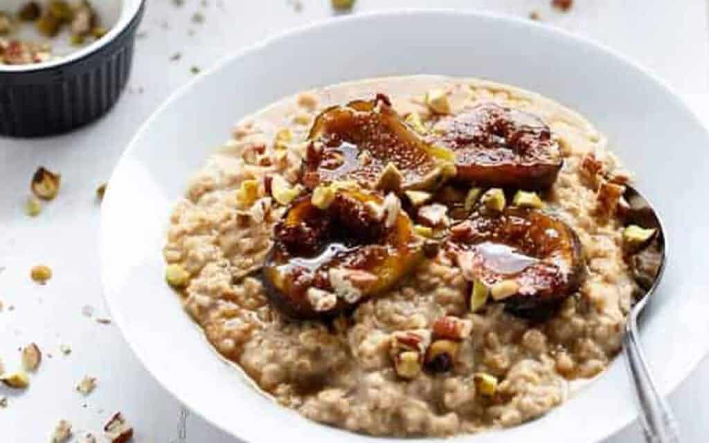 a delicious full bowl of porridge with caramelized figs on top and a spoon on the side, the table has a few scattered crushed pistachios.