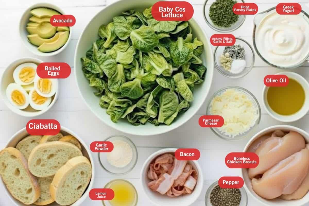 Flat lay of ingredient shot of avocado, boiled eggs, baby cos lettuce, dried parsley flakes, greek yogurt, anchovy fillets, garlic and salt, olive oil, parmesan cheese, boneless chicken breasts, pepper, bacon, ciabatta, garlic powder, lemon juice