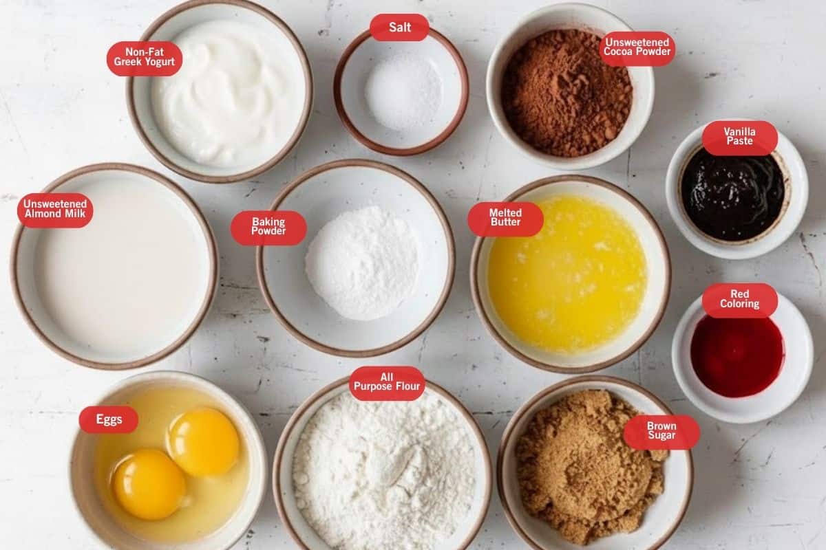 Flat lay photo of ingredient shot of non fat greek yogurt, salt, unsweetened cocoa powder, vanilla paste, red food coloring, brown sugar, all purpose. flour, eggs, melted powder, baking powder, unsweetened almond milk