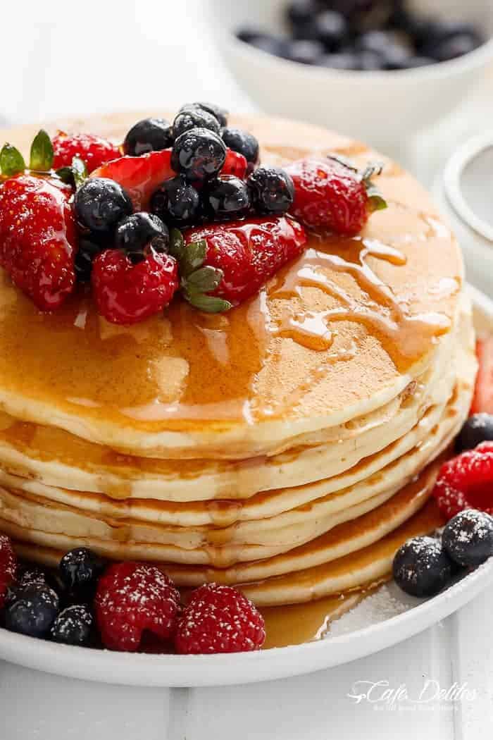Close-up of a stack of pancakes drizzled with maple syrup and garnished with mixed-berries, cascading from the top to the bottom of the stack.