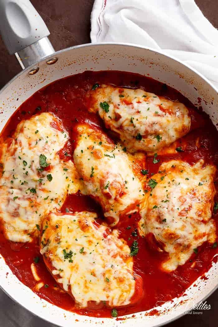 Five chicken breasts pieces, covered in grilled cheese, sprinkled with herbs, on a bed of tomato sauce in a white pan.