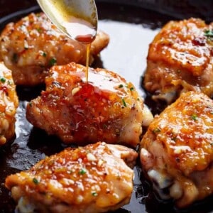 This close-up image of pan-seared chicken thighs coated in a vibrant orange glaze being drizzled with more of the glossy sauce from a spoon.