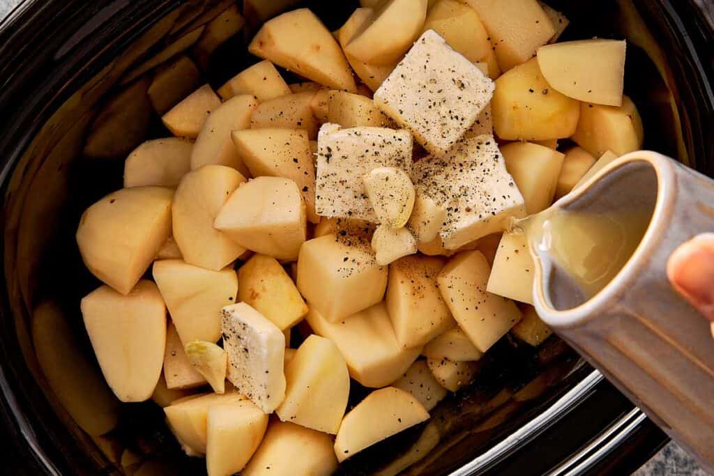 a top down view of cut up in cubes white potatoes in a slow cooker with butter cubs inside and a jug of milk being poured in