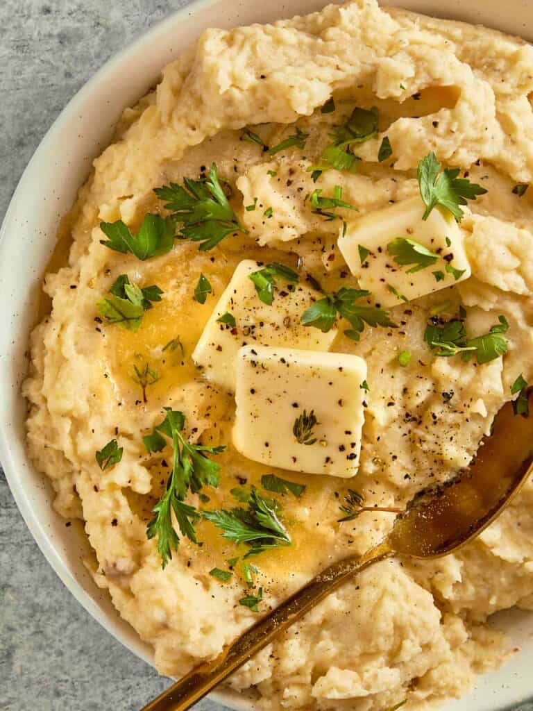 top down view of a white bowl filled with creamy potatoes and two cubes of butter with herbs on top close up shot with chives on top
