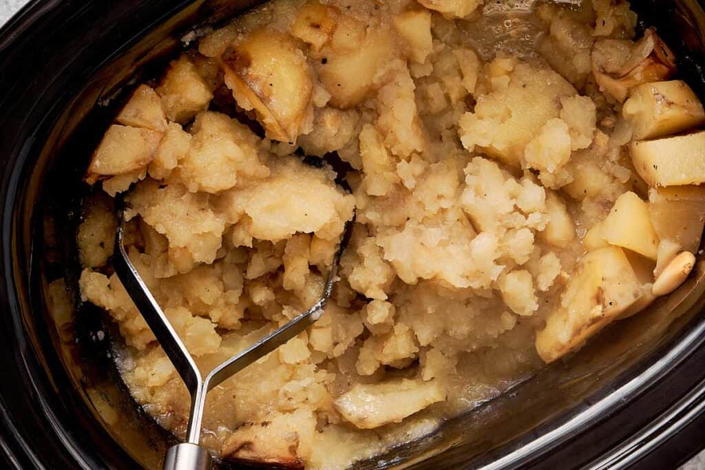 a top down view of baby potatoes in a slow cooker being mashed with a potato masher