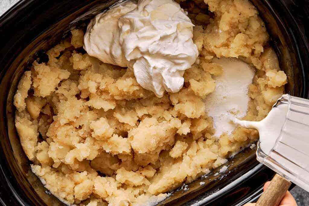 a top down shot of creamy potaotes already mashed and a dollop of cream on top and pouring milk in a slow cooker