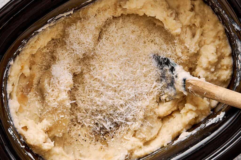 a top down view of mashed potatoes very creamy stirred by a wooden spoon in a slow cooker with parmesan cheese on top