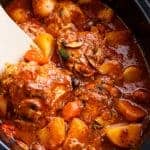 A rich, red stew full of chicken, roughly chopped vegetables being stirred by a white spatular in a slow cooker.