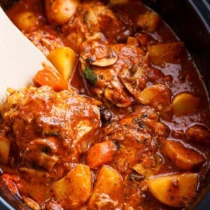 A rich, red stew full of chicken, roughly chopped vegetables being stirred by a white spatular in a slow cooker.