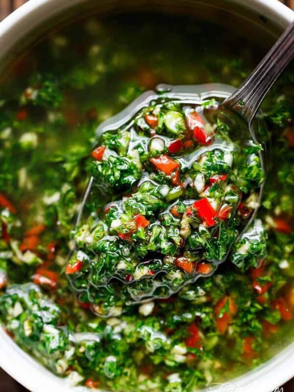 A small serving spoon lifts a spoonful of vibrant chimichurri, showcasing green herbs and red chili pieces mixed with oil from a bowl.