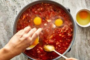 a top down view of a pan filled with diced vegetables and thick tomato salsa with two egg yokes raw cooking in the top and a spoon mixing with one hand and the other hand is adding in a bowl of sauce