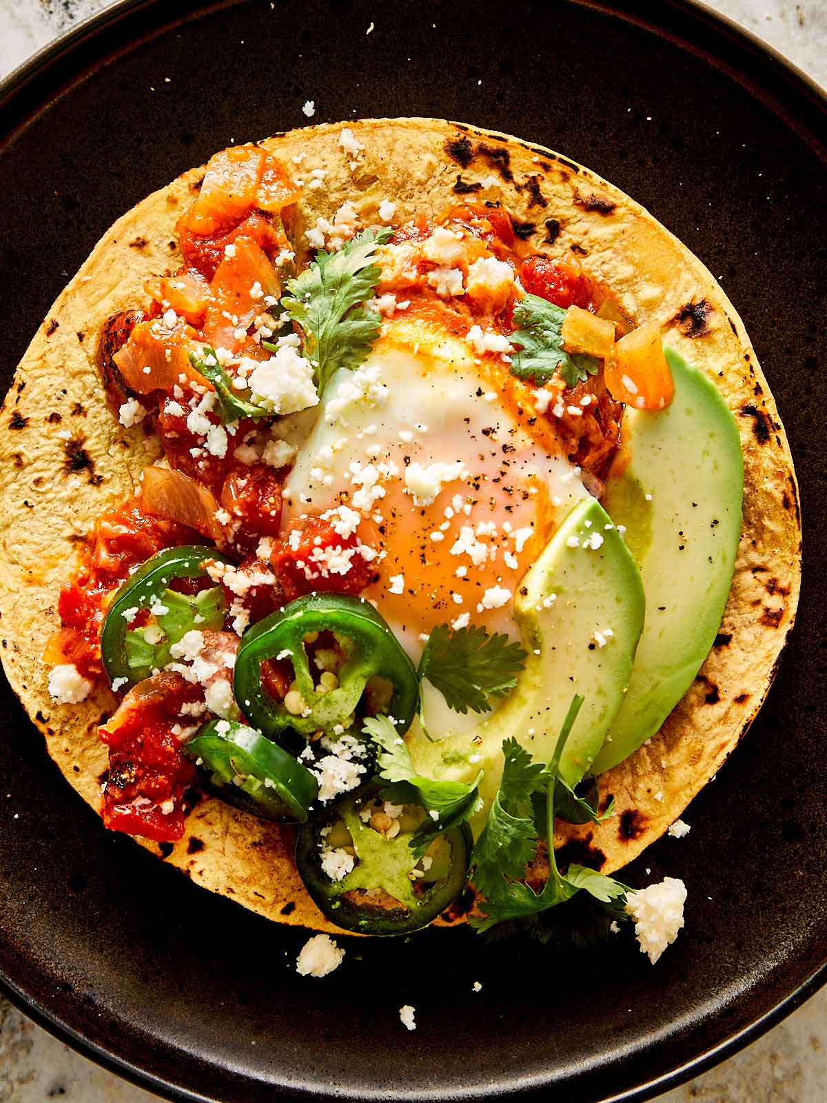 a top down view of a black plate with a large corn tortilla with eggs and tomato salsa and avocado in the middle