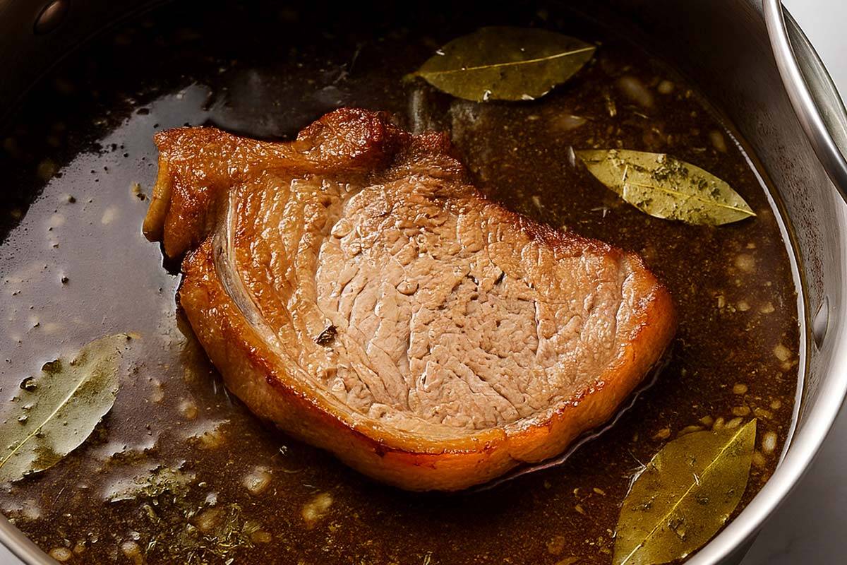 Pork shoulder cooking in a slow cooker, lightly browned on top, surrounded by bay leaves and a rich, seasoned broth.
