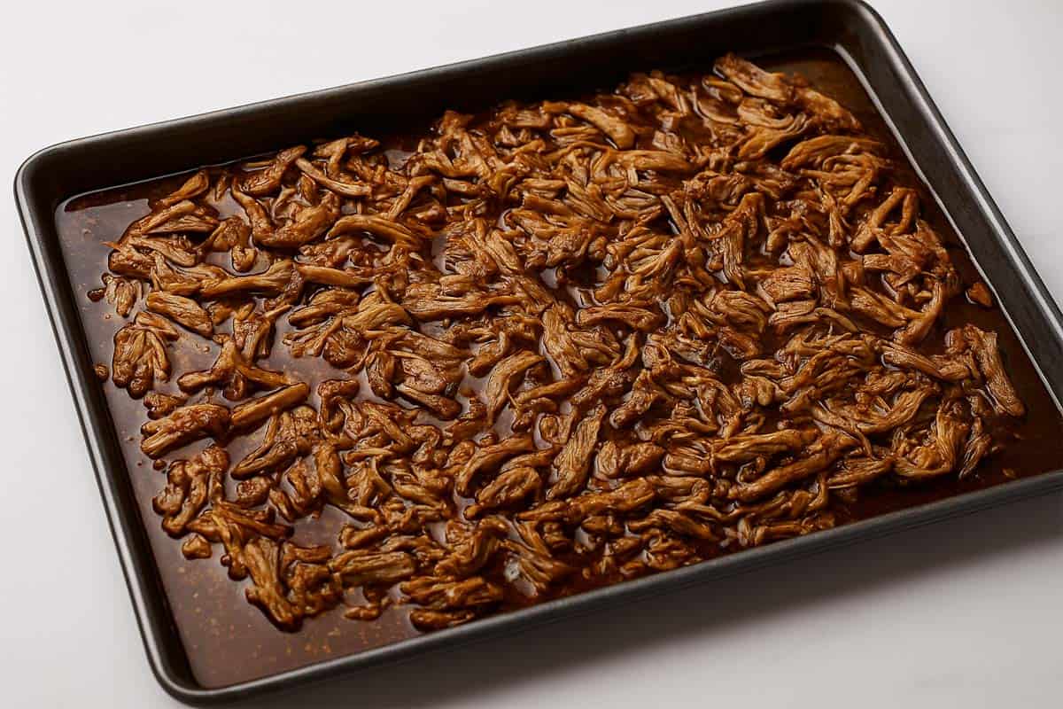 Crispy shredded pork spread evenly on a baking sheet, drizzled with cooking liquid, ready to broil until golden.