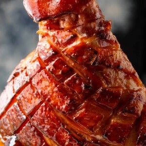 This close-up shot of beautifully glazed ham, its surface glistening with a rich, caramelized coating. The ham has been meticulously scored in a diamond pattern, allowing the glaze to penetrate the meat and create tantalizing crevices of flavor. The deep, reddish-brown hue suggests a savory and slightly sweet profile. A generous amount of glaze pools around the base, inviting you to savor its deliciousness. The background is a dark gray, providing a neutral canvas that accentuates the ham's visual appeal.