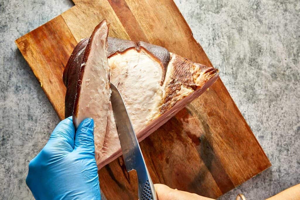 A top down view of a leg of ham on a wooden chopping board with a knife removing the rind off the leg ham exposing a white layer of fat