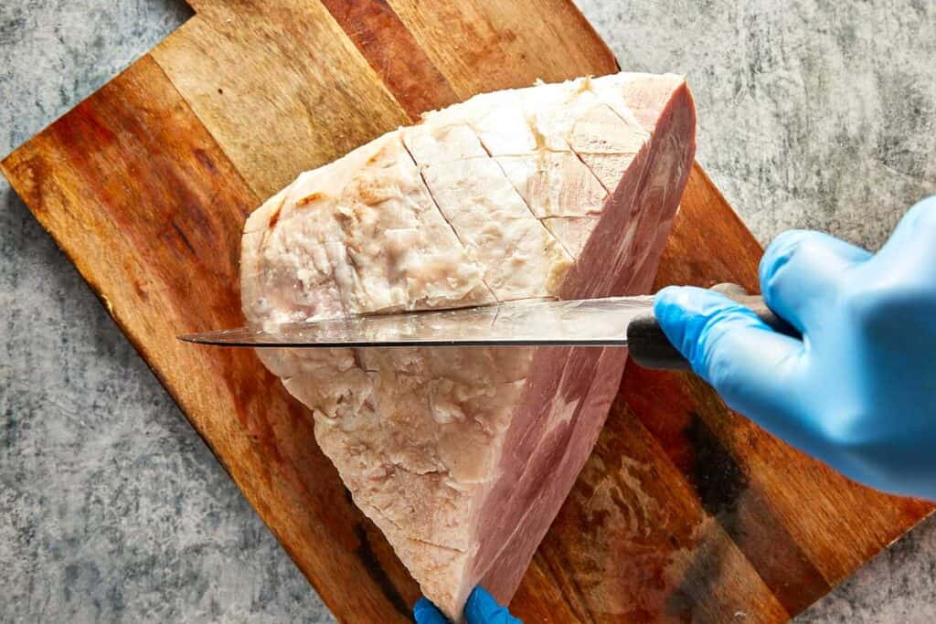 a knife cutting the ham into a diamond shape on a wooden board sitting on top of kitchen bench
