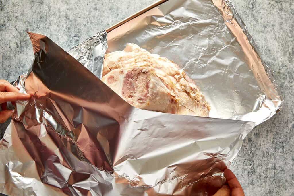 a top down view of a leg ham sitting on aluminum foil, the leg is sitting cut side down looking like a mountain from top down and exposing the fatty layer and foil covering the top of the ham