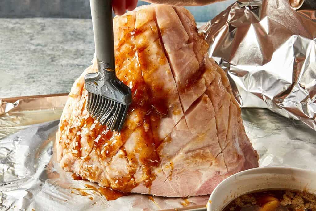 a side view of leg ham with a brush coating it with a glaze and a bowl of glaze on the side.