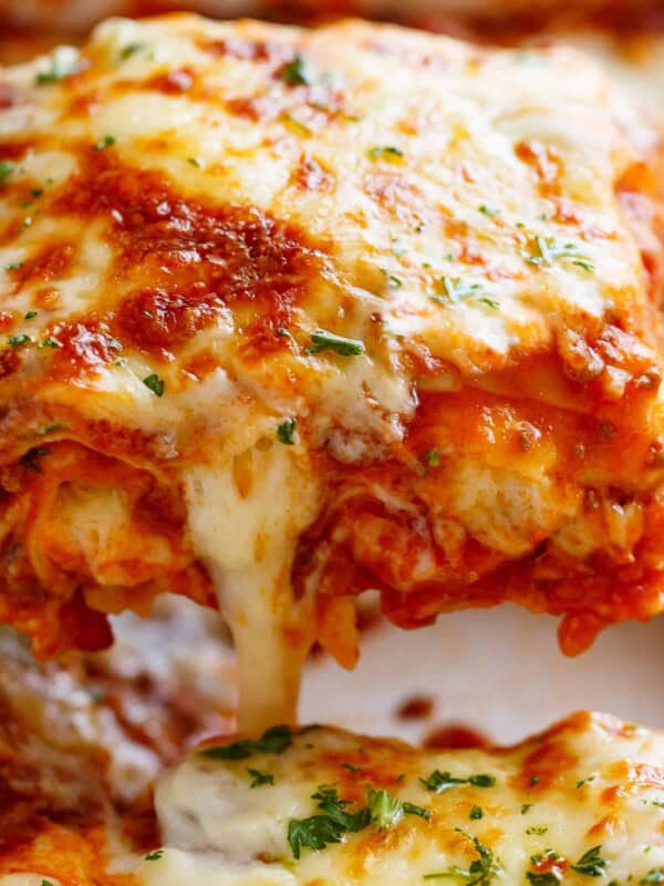 A beautiful close-up of a slice of lasagna being lifted by a spatula from the baking tray, showcasing layers of pasta, rich meat sauce, creamy cheese, and a golden, bubbly top.