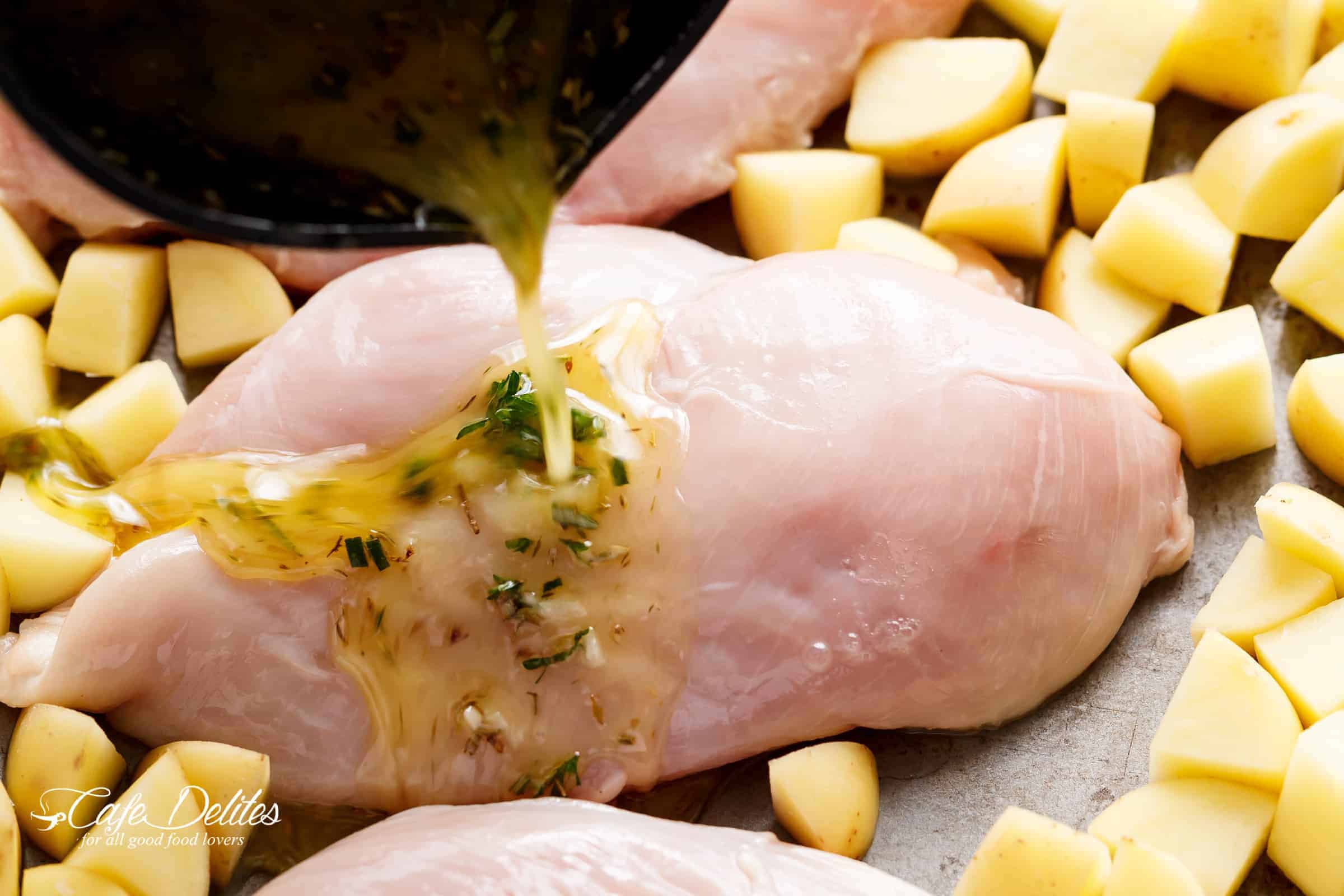 raw chicken breast piece is surrounded by uncooked potatoes on a sheet pan with garlic sauce being poured over top.