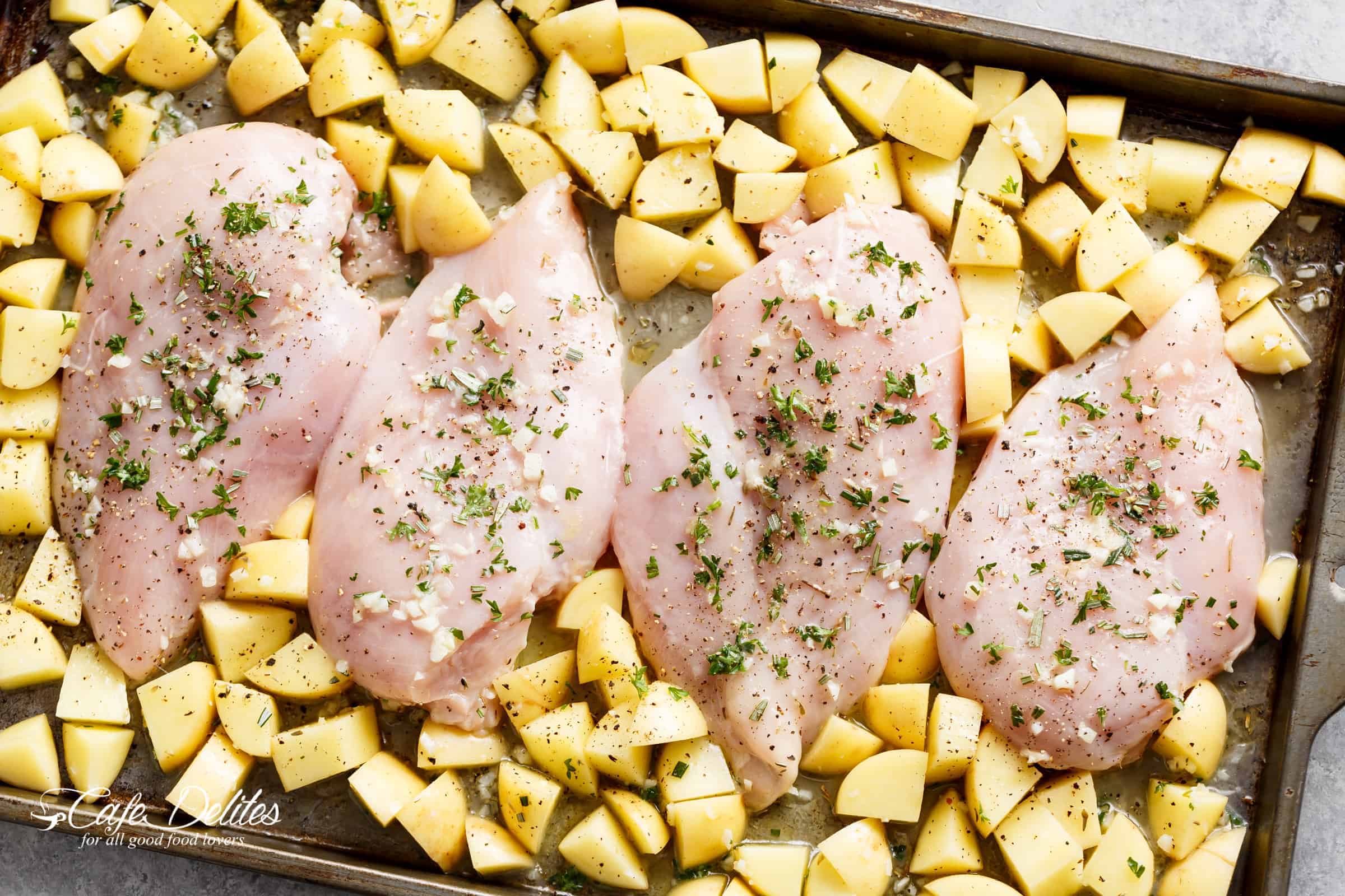 Four raw chicken breasts pieces covered in garlic sauce and herbs on a tray with cubed potato.