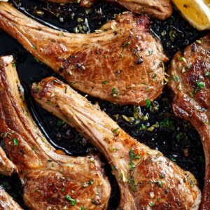 cooked lamb chops pieces in a black pan, green herbs sprinkled on top with two lemon wedges to one side.