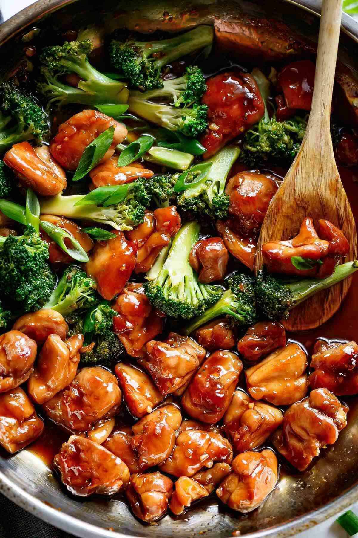 Close-up image of Teriyaki Chicken with broccoli and chives in a pan, being stirred by a wooden spoon soaked in teriyaki sauce.