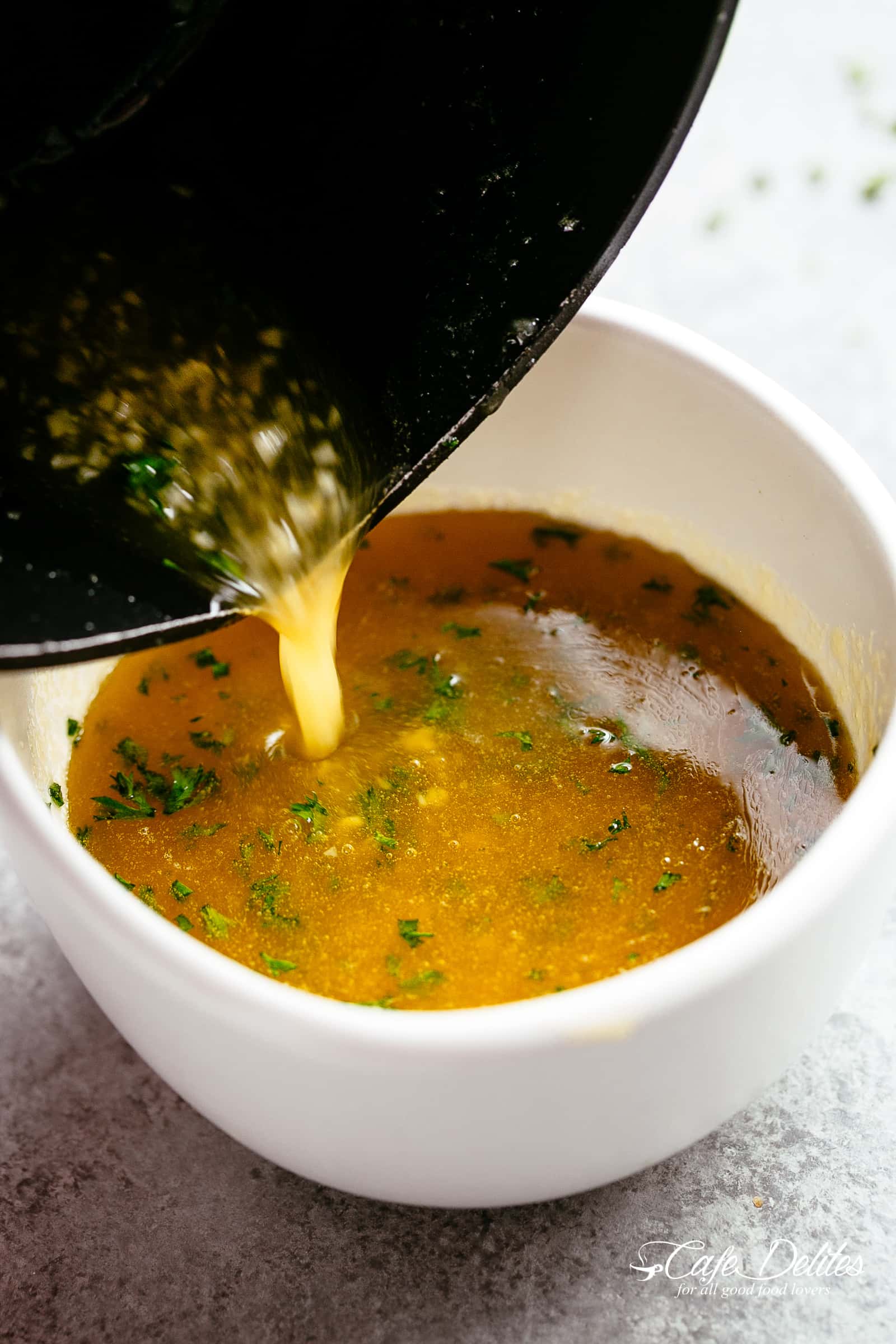 How To Make Honey Garlic Butter | cafedelites.com Hot & Spicy Golden Honey Garlic Butter Sauce pouring from a black pot into small white sauce cup with small bits of garlic and parsley.