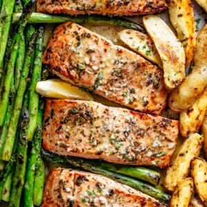 The image shows four salmon fillets on a baking sheet, along with asparagus spears and potato wedges. The salmon is covered in a brown glaze and garnished with fresh herbs and lemon slices. The bright green baked asparagus is soft and the potatoes are slightly browned after baked