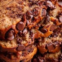 freshly baked banana bread has a golden brown crust and the inside is a pale yellow color. The bread is studded with chocolate chips that have melted slightly, creating a gooey texture within the slices. The camera is zoomed into a stack of these slices, emphasizing the moist texture and abundance of chocolate chips within each piece.