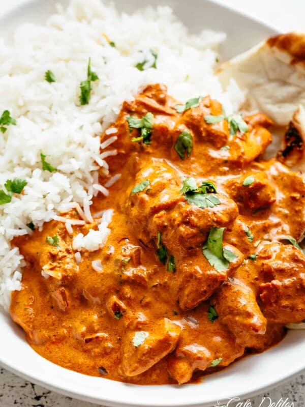 A close-up shot of Butter Chicken, generously garnished with fresh coriander, alongside a bed of fluffy white rice and a piece of naan bread. Chunks of chicken, coated in the flavorful sauce, are scattered throughout the dish.