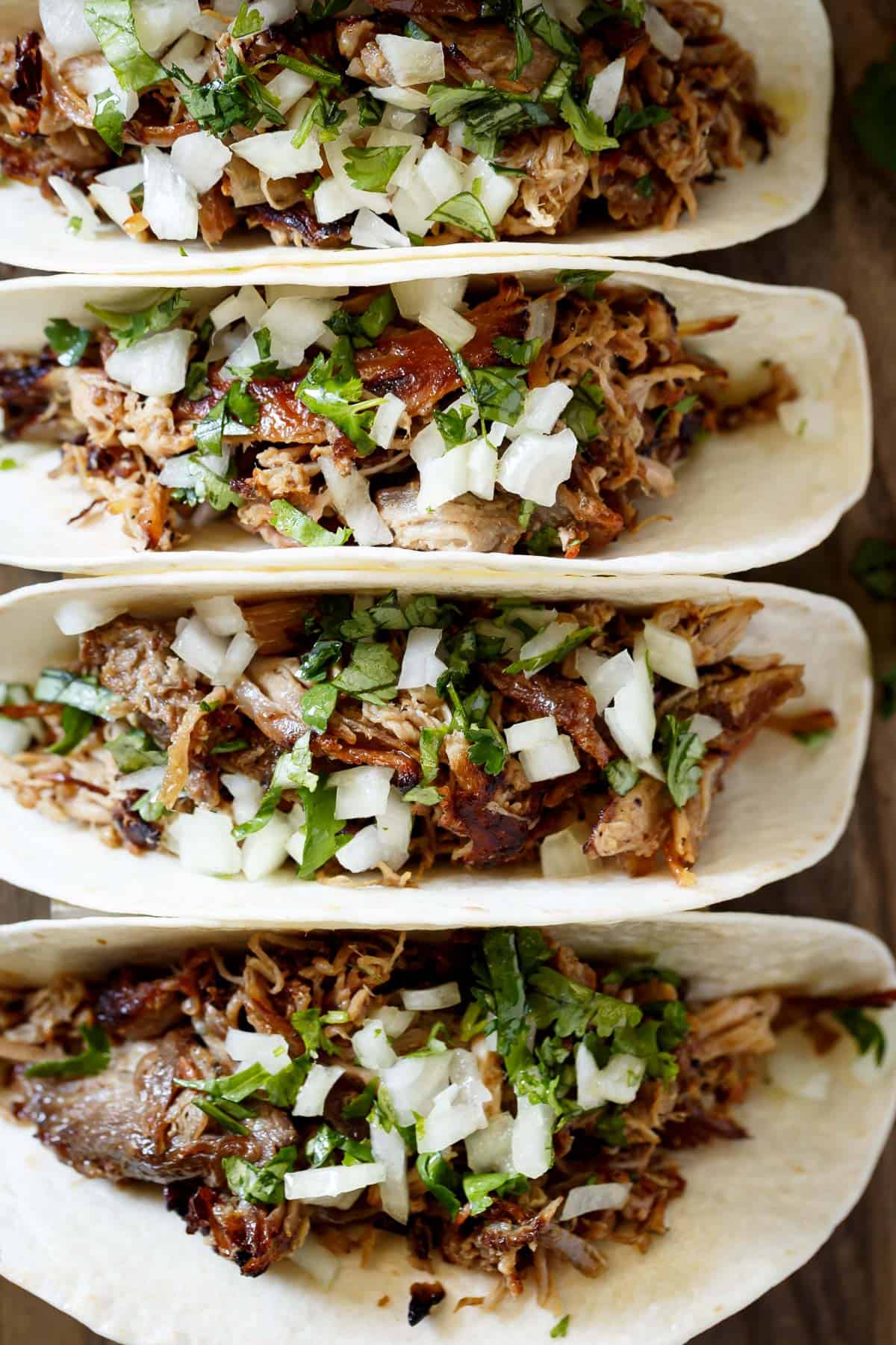 The image shows four corn tortillas, each filled with carnitas, chopped white onion, and chopped cilantro. The tortillas are laid out in a row, slightly overlapping, on a wooden table or cutting board. 