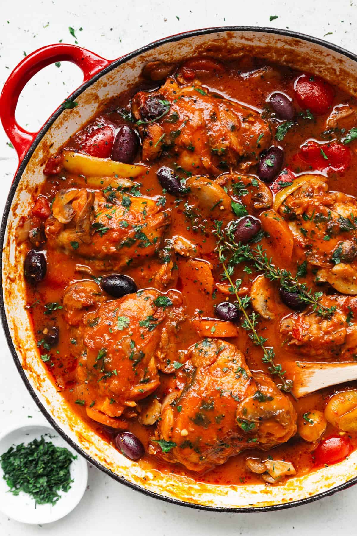 Spicy Five chicken pieces in a le creuset style casserole dish covered in red Chicken Cacciatore sauce. Olives, carrots and other vegetables are submerged in the sauce.