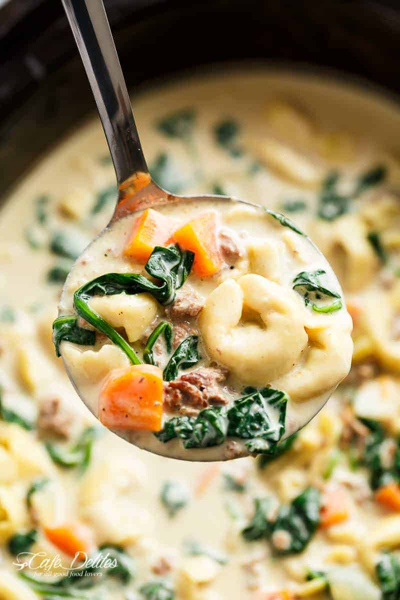 Close-up of a ladle lifting a portion of the soup, showcasing tender tortellini, vibrant spinach leaves, hearty Italian sausage, and chunks of carrot before serving it into a bowl.