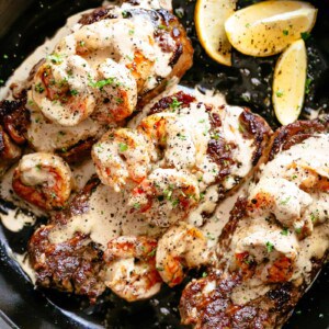 Three pieces of cooked steak in a pan with prawns laid on top and covered in creamy garlic sauce.
