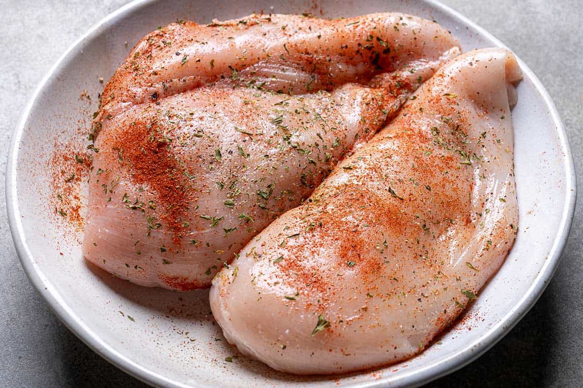 Chicken breasts in a white bowl, evenly coated with salt, pepper, paprika, dried parsley, and 2 teaspoons of oil, ready for searing.