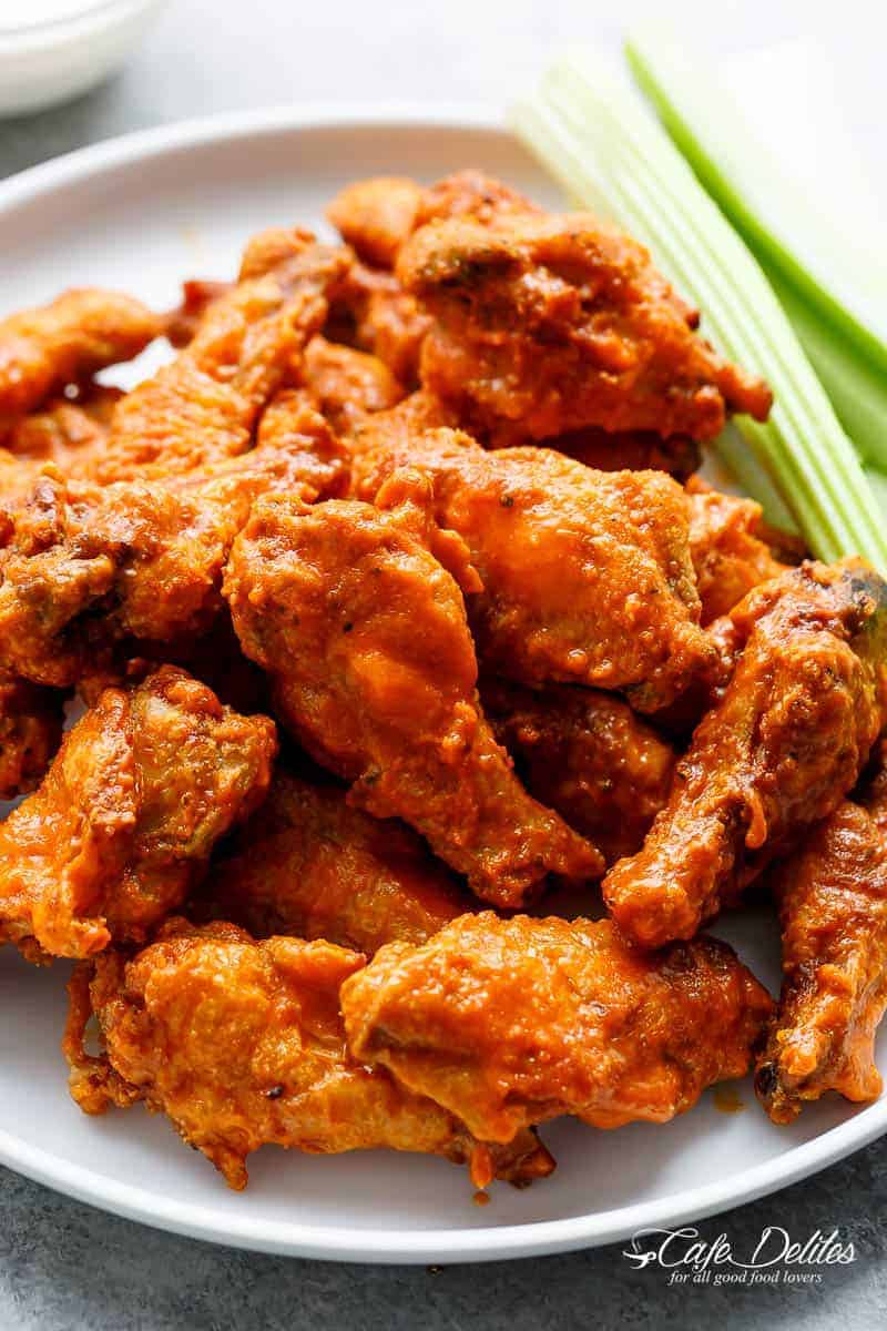The image showcases a delicious pile of chicken wings coated in a vibrant orange buffalo sauce. The wings are piled high on a white plate, their glistening skin reflecting light and hinting at their crispy texture. Accompanying the wings are several sticks of fresh celery, providing a cool and crunchy contrast to the spicy wings. The background is intentionally blurred, further emphasizing the chicken wings as the star of the show.