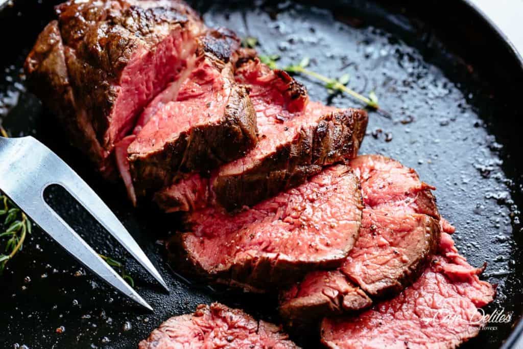Roasted Beef  in a black skillet cut into slices cooked to medium rare with sprigs of fresh rosemary and a crack of pepper over the meat.