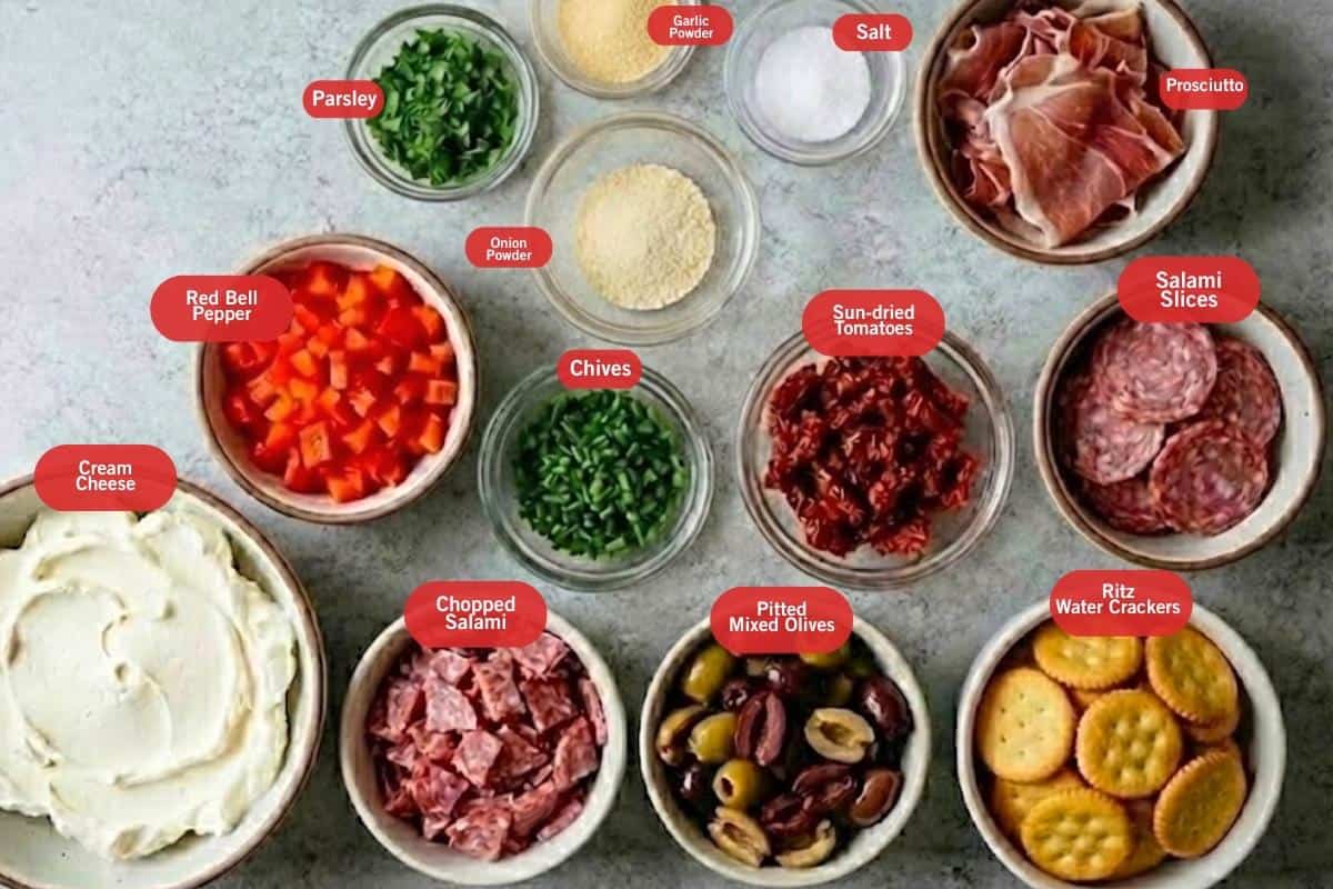 Flat lay photo of ingredient shot of parsley, onion and garlic powder, salt, prosciutto, salami slices, ritz water crackers, pitted mixed olives, chopped salami, cream cheese, red bell pepper, chives, chopped sun-dried tomatoes