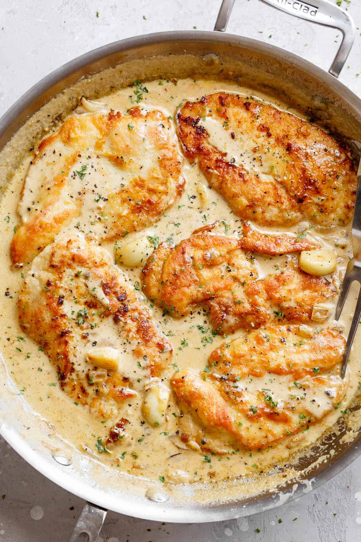 A silver frying pan of deliciously five Chicken Breasts in a garlic cream sauce with whole garlic cloves and a silver serving fork 