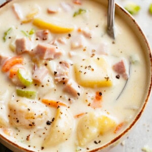 creamy potato soup with a silver spoon resting in the top right corner. The bowl is on a marble bench top, a grey kitchen towel to the left of the bowl, and green celery bits scattered near the top of the frame.The soup has diced ham, potatoes, carrots, and is topped with pepper.
