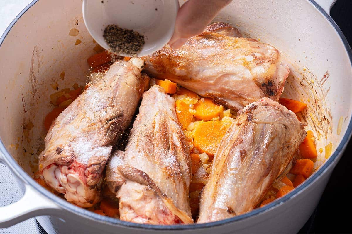carrots, diced onion and lamb shanks in a blue pot being sprinkled with salt and pepper