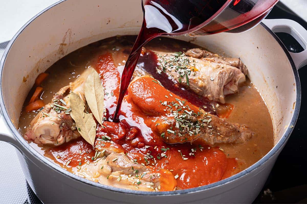 carrots, diced onion and lamb shanks and stock in a blue pot with wine being poured in