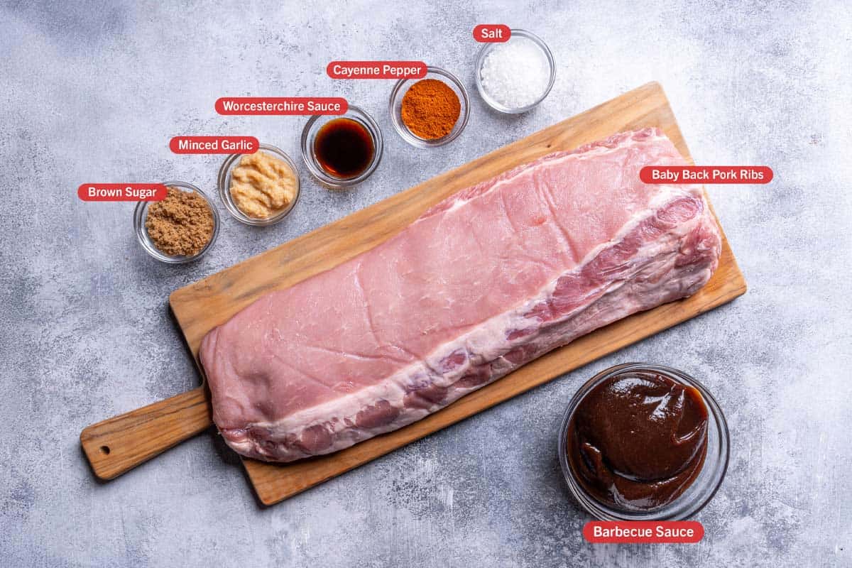 Ingredients for slow cooker barbecue ribs laid out on a grey textured background, including brown sugar, minced garlic, Worcestershire sauce, cayenne pepper, salt, baby back pork ribs, and barbecue sauce, all labeled.
