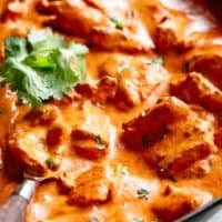 A close-up shot showcases a pan filled with a vibrant, creamy tomato-based curry. Large chunks of chicken are coated in the rich sauce, and fresh cilantro sprigs are sprinkled throughout. A wooden spoon rests in the bottom left corner of the pan, ready to serve. The background is slightly blurred, drawing the focus to the appetising curry.