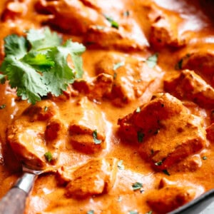 A close-up shot showcases a pan filled with a vibrant, creamy tomato-based curry. Large chunks of chicken are coated in the rich sauce, and fresh cilantro sprigs are sprinkled throughout. A wooden spoon rests in the bottom left corner of the pan, ready to serve. The background is slightly blurred, drawing the focus to the appetising curry.