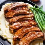 Slow-cooked barbecue ribs resting on a bed of mashed potatoes, accompanied by a side of green beans, served on a black serving plate.